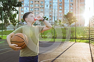 A teenager drinks water, on an open playground with a basketball ball. Healthy lifestyle and sport concepts