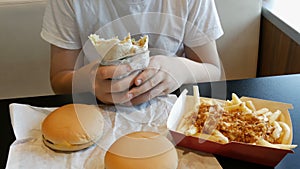 Teenager eats a big chicken roll in a fast food cafe. On the table are large cheeseburgers and French fries.
