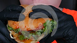 A teenager eats big juicy hamburger with an egg, a tomato cutlet, salad and red sauce. Hands in special black gloves