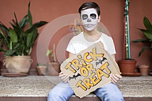 Teenager at the entrance of his house. He has his face made up and holds a sign with the text Trick or Treat