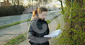 Teenager female booklover read book outdoors standing on sidewalk at city road.