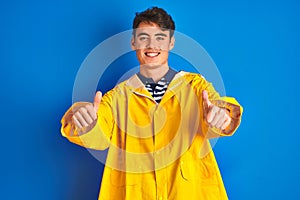Teenager fisherman boy wearing yellow raincoat over  background approving doing positive gesture with hand, thumbs up