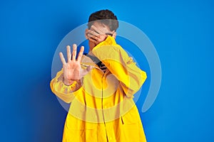 Teenager fisherman boy wearing yellow raincoat over isolated background covering eyes with hands and doing stop gesture with sad