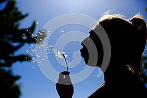 The teenager gently blows and spreads the dandelion seeds against the blue sky.