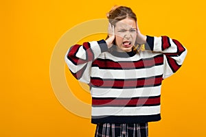 Teenager girl in a casual striped sweater asks to be quieter covering her ears on an orange background with copy space