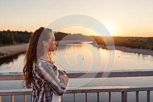 teenager girl with curly hair in lifestyle clothes standing near a railing on the bridge looking at sunrise