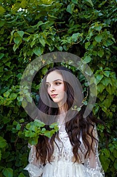 Teenager girl with curly long hair in white dress on green ivy background.