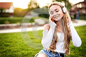 Teenager girl enjoying life and sitting in the grass in a public park, while listening to music with headphones on her head with
