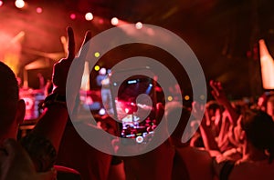 Teenager girl fan raised hand with V sign Gesture and shooting vertical video with smartphone popular band on the night concert.