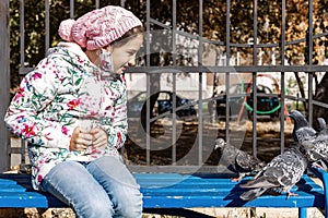 Teenager girl feeding pigeons in the park. The girl sits on a bench and feeds the birds with seeds. Autumn sunny day