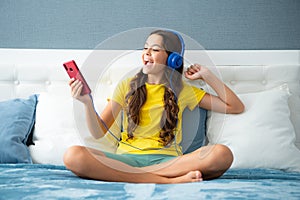 Teenager girl in headphones relax on bed at home using phone. Child in earphones browse internet on smartphone