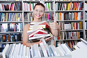 teenager girl holding stack of books shows thump up in a bookstore