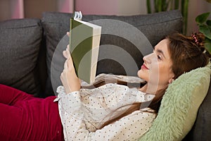 Teenager girl home - student read book laying on sofa. Portrait of smiling woman holding book and lying on couch.