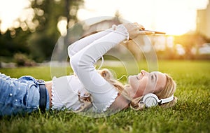 Teenager girl lies in grass in a park and listening to music on headphones while holding her smartphone overhead and typing on the