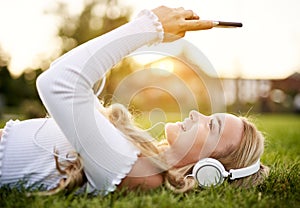 Teenager girl lies in grass in a park and listening to music on headphones while holding her smartphone overhead and typing on the