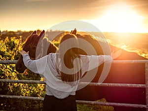 Teenager girl with long hair by a metal gate to a field with dark horses. Selective focus. Models hands on horses faces.