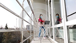 Teenager girl in mask and headphones, listening to music on laptop, chatting online, on open balcony. spring sunny day