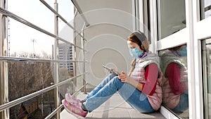 Teenager girl in mask and headphones, listening to music on tablet, on open balcony. spring sunny day. quarantine. stay