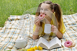 Teenager girl in nature, lies on a plaid, eats donuts and reads a book. In the hands of a white mug with a tasty drink