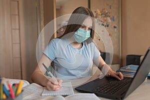 Teenager girl protective medical mask, writes notebook, security virus infections, electronic education at home, laptop