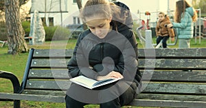 Teenager girl read book while sitting on bench on kids playground on autumn day.