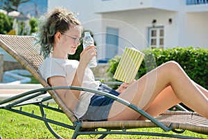 Teenager girl resting lying in an outdoor chair on lawn, reading book, drinks water from bottle