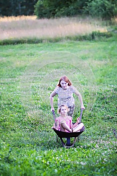 Teenager girl rides her sister in the old iron wheelbarrow in the village at summer