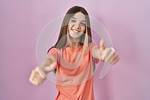 Teenager girl standing over pink background approving doing positive gesture with hand, thumbs up smiling and happy for success