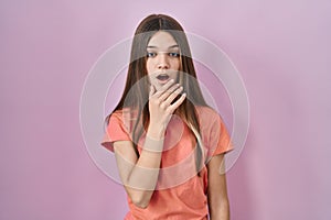 Teenager girl standing over pink background looking fascinated with disbelief, surprise and amazed expression with hands on chin