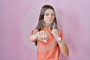 Teenager girl standing over pink background punching fist to fight, aggressive and angry attack, threat and violence