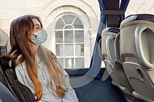 Teenager girl traveling on a public bus sitting by a window and wearing blue surgical mask. Selective focus. The model has long