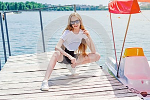 Teenager girl in a white T-shirt and sunglasses on a small wooden pier on the lake shore. Sitting doing stretching