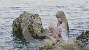 Teenager girl in white tunic sitting on rocks in sea water on summer beach. Woman photographer in bikini photographing