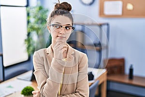 Teenager girl working at the office wearing glasses serious face thinking about question with hand on chin, thoughtful about