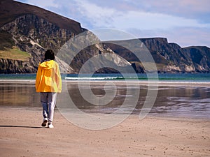 Teenager girl in yellow jacket walking on a wet sand of a beach, beautiful nature scenery in the background. Selective focus. Keel