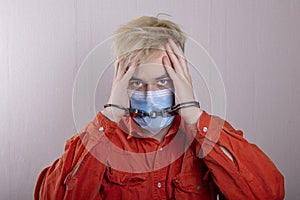 A teenager in handcuffs and a medical mask holds his head against a gray background.Headache for juvenile delinquents in quarantin