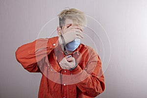 A teenager in handcuffs and a medical mask holds his head against a gray background.Headache for juvenile delinquents in quarantin