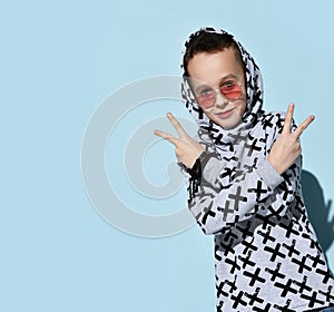 Teenager with headphones on neck, in sunglasses, hoodie, black bracelet. Smiling, showing victory hands, posing on blue background