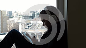 Teenager with headphones sitting on window sill and enjoy listening to music from his cell phone at home