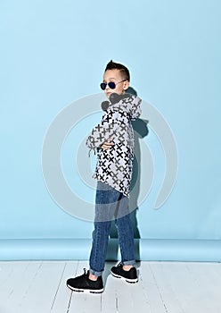 Teenager in headphones, sunglasses, casual clothes. Looking wondered, listening to music. Sitting on white chair. Blue background