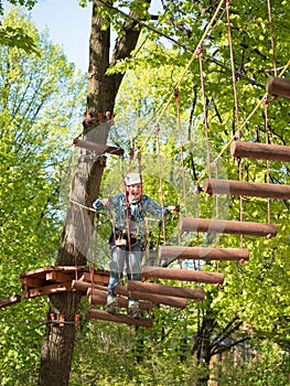 Teenager in helmet and with a safety rope boy goes on suspension bridge made of logs on the blurred background