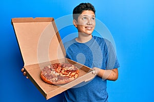 Teenager hispanic boy eating tasty pepperoni pizza smiling looking to the side and staring away thinking