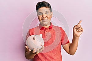 Teenager hispanic boy holding piggy bank smiling happy pointing with hand and finger to the side