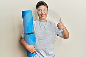 Teenager hispanic boy holding yoga mat smiling happy and positive, thumb up doing excellent and approval sign