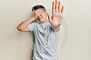 Teenager hispanic boy wearing casual grey t shirt covering eyes with hands and doing stop gesture with sad and fear expression