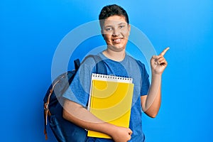 Teenager hispanic boy wearing school bag and holding books smiling happy pointing with hand and finger to the side