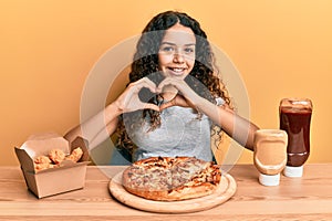 Teenager hispanic girl eating pizza and fried chicken smiling in love doing heart symbol shape with hands