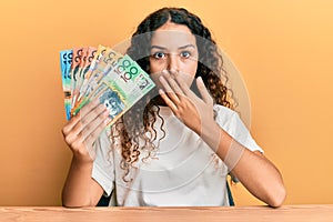 Teenager hispanic girl holding australian dollars covering mouth with hand, shocked and afraid for mistake