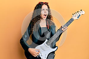 Teenager hispanic girl playing electric guitar angry and mad screaming frustrated and furious, shouting with anger