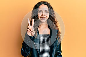 Teenager hispanic girl wearing black leather jacket smiling with happy face winking at the camera doing victory sign